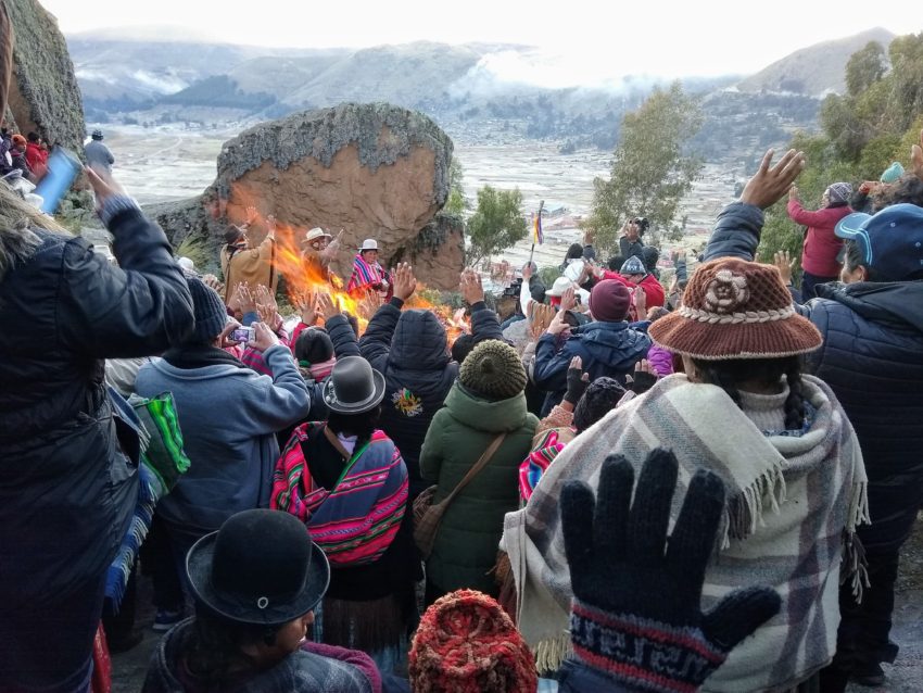 Many people facing away from the camera, with hands raised, facing a fire and the coming sunrise on the Winter Solstice at Lake Titikaka in Bolivia.