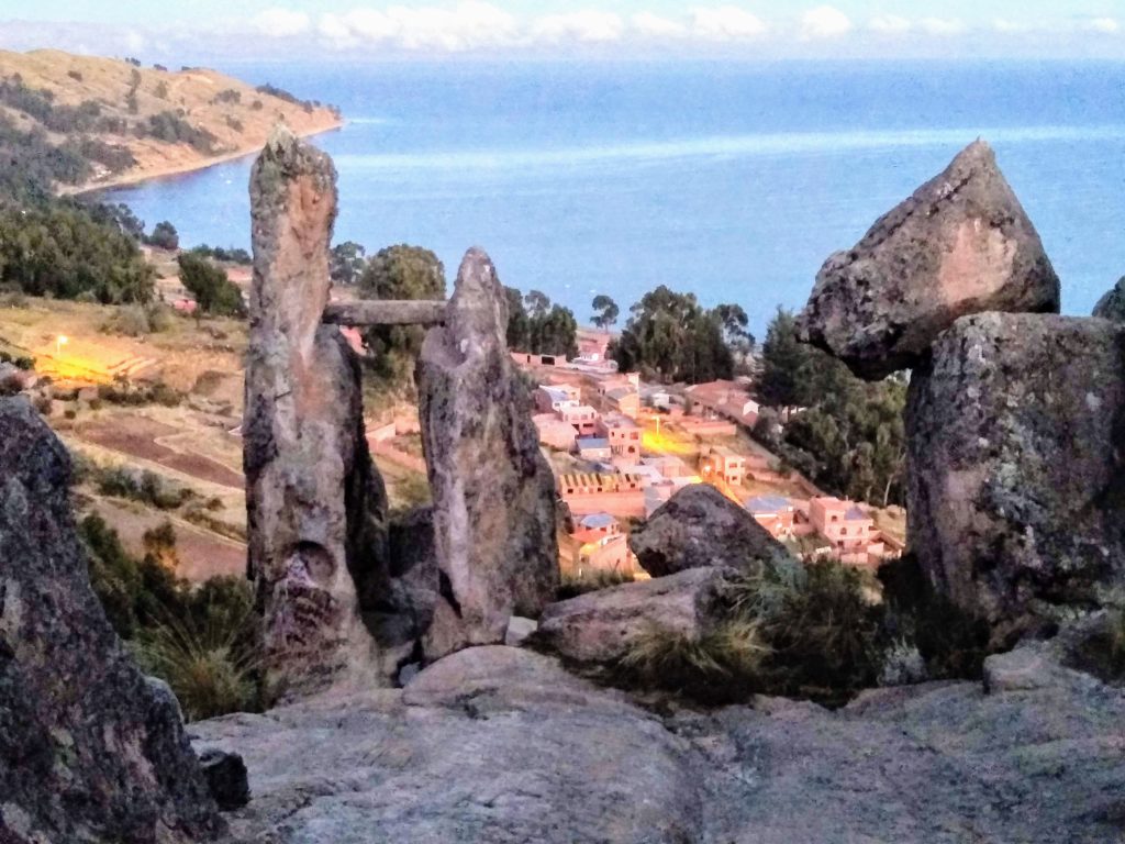 Two tall pillars of stone supporting a horizontal stone beam surrounded by rock outcroppings and overlooking a blue lake.