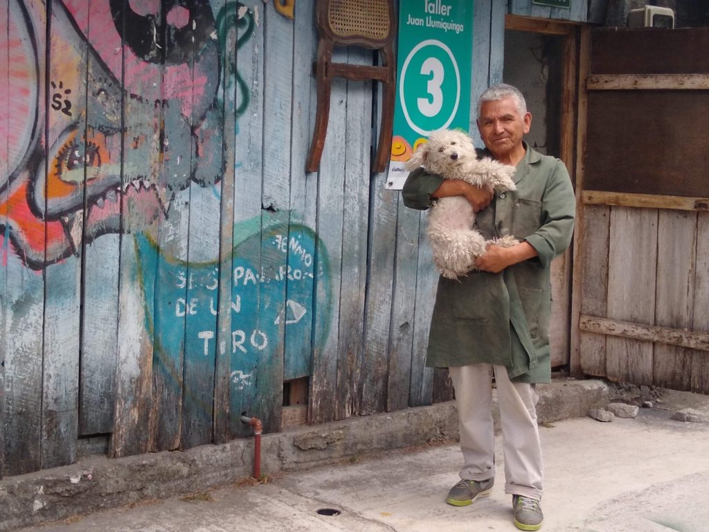 An older man holding a small white dog standing next to a mural painted on the front of the man's house.