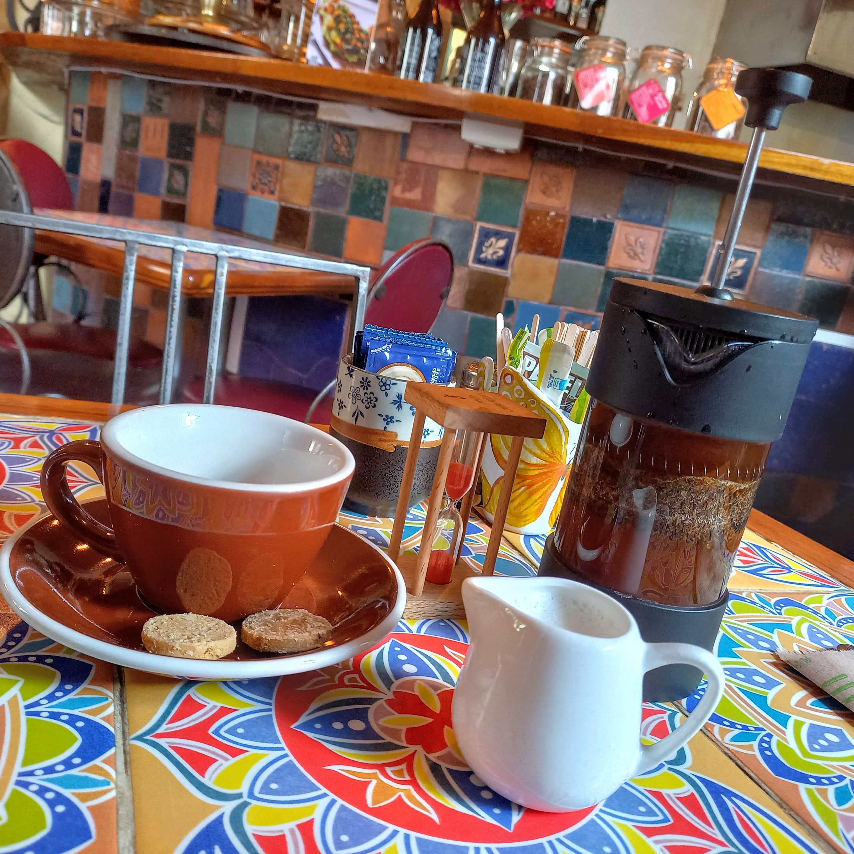 brown coffee cup next to a french press filled with black coffee and a white cream container on a colorful table cloth with colorful tile in the background